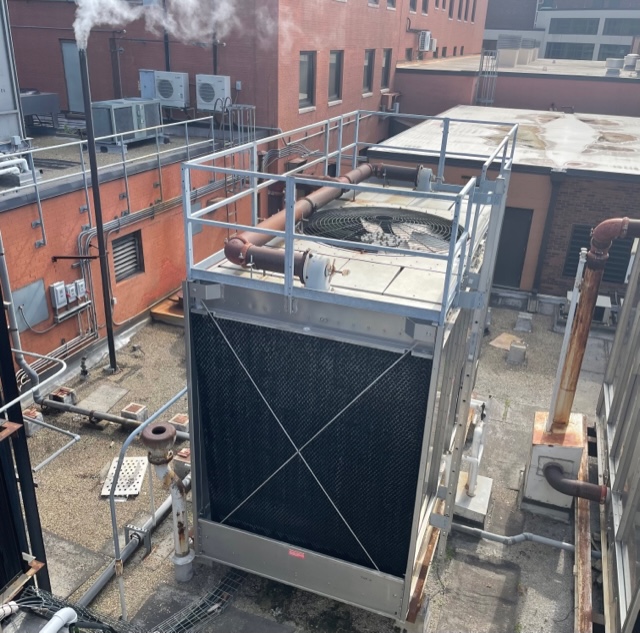A brick and concrete rooftop view of a metal cooling tower and other HVAC equipment.
